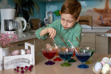 A beautiful sweet and funny red -haired boy paints eggs in the kitchen for Easter. Multi -colored eggs, the child paints at home