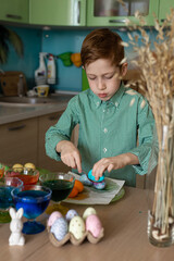 A beautiful sweet and funny red -haired boy paints eggs in the kitchen for Easter. Multi -colored eggs, the child paints at home. Close -up portrait