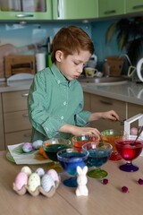 A beautiful sweet and funny red -haired boy paints eggs in the kitchen for Easter. Multi -colored eggs, the child paints at home, portrait