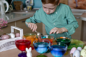 A beautiful sweet and funny red -haired boy paints eggs in the kitchen for Easter. Colored eggs, the child paints at home. Close -up