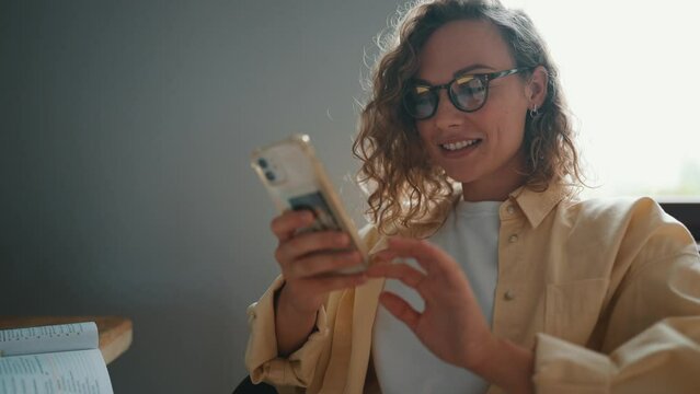 Curly haired smiling woman typing on mobile while sitting at the table at home