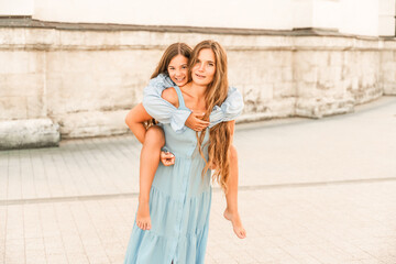 Mother of the daughter walks playing. Mother holds the girl on her back, holding her legs, and her daughter hugs her by the shoulders. Dressed in blue dresses.
