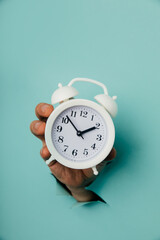 Hand holds alarm clock through a blue paper hole. Time management and deadline concept