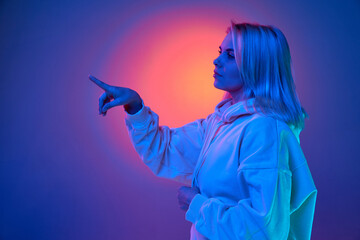 Portrait of young beautiful woman pointing with finger over gradient purple studio background in blue neon light. Concept of emotions, facial expression, lifestyle, inspiration, sales, ad