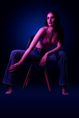 Fototapeta na wymiar Beautiful young caucasian brunnete girl sitting on chair in sexy lingerie and jeans in colored blue purple light in studio on a dark background
