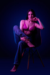 Obraz na płótnie Canvas Beautiful young caucasian brunnete girl sitting on chair in sexy lingerie and jeans in colored blue purple light in studio on a dark background