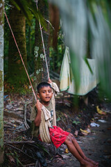 Little Bangladeshi kid playing with a swing made with clothes and rope using trees as support in a village garden 