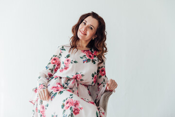 a brunette woman in a floral dress sits in a chair on a light background