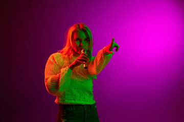 Relaxation and hobby. Young woman singing in microphone over magenta studio background in green neon light. Concept of emotions, facial expression, lifestyle, inspiration, sales, ad