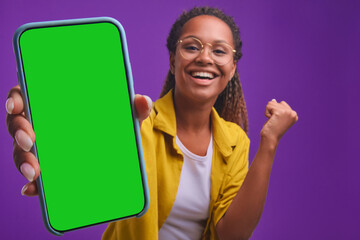 Young overjoyed African American woman stretches out hand with smartphone showing off record in mobile game or result in application with intelligence test stands in purple studio. phone green screen - obrazy, fototapety, plakaty