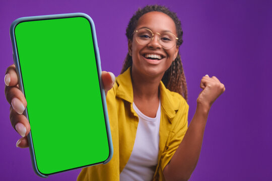 Young overjoyed African American woman stretches out hand with smartphone showing off record in mobile game or result in application with intelligence test stands in purple studio. phone green screen