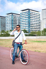 Obraz premium Man with sunglasses standing with his bicycle talking by phone and the city in the background