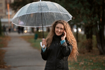 Laughing beautiful girl in autumn park keeps transparent umbrella talking on phone. Young woman walking in the park