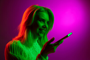 Happy smiling young woman talking on mobile phone, recording voice messages on magenta studio background in green neon light. Concept of emotions, facial expression, lifestyle, inspiration, sales, ad