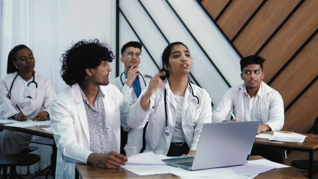 Young medical interns of mixed race in medical clothing studying together human anatomy at lectures of surgery in light office in hospital, asking questions to professor, raising hand 