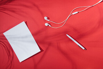 Top view of tropical red background with pen, headphones, and creative copyspace with paper frame. Minimal summer concept. Flat lay. Palm tree leaf shadow.