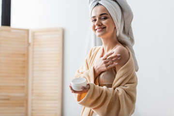 pleased young woman with towel on head holding container while applying body butter.