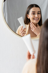 happy young woman in bathrobe holding tube with cream and looking at mirror in bathroom.