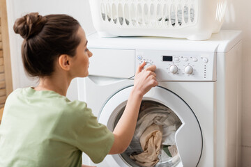 Side view of blurred woman switching washing machine near basket in laundry room. - obrazy, fototapety, plakaty