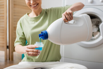 Cropped view of smiling woman pouring liquid washer in laundry room.