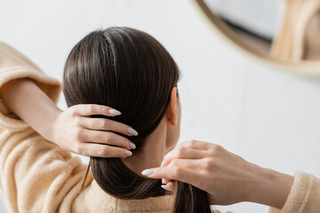 Naklejka premium back view of young brunette woman adjusting shiny hair near mirror in bathroom.