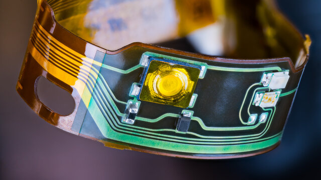 Flex printed circuit and small electronic components in plastic strip curled into circle on dark background. Closeup of ribbon cable with green and yellow copper lines on flexible PCB from headphones.