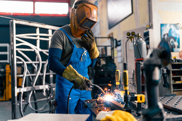 A worker is using a welding machine in his hand and working on a metal rod, he is wearing his visor for protection