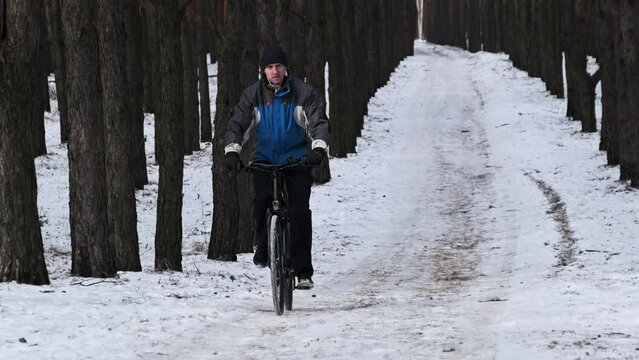 Man ride a bicycle along a snowy path between winter trees in slow motion. Young man biking follows the forest track in woodland. Cyclist travel on bike. Healthy lifestyle, outdoor walking, sport