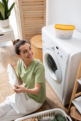 High angle view of smiling woman looking at box on washing machine near basket with clothes in laundry room.