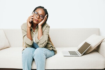Stylish African American woman business freelancer working sitting on the couch at home in a laptop and phone, business calls and correspondence, home clothes and eyeglasses, light interior background