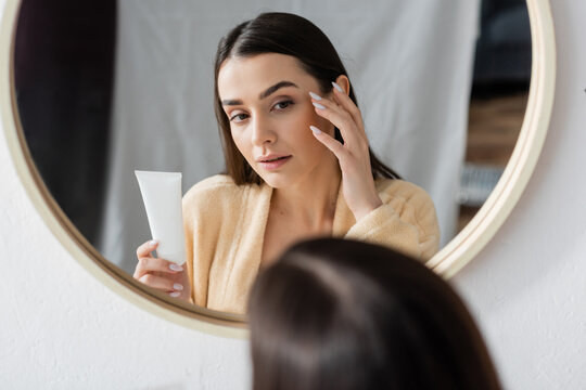brunette young woman in bathrobe holding tube with cream and looking at mirror in bathroom.