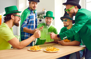 Group of happy male friends celebrating St Patrick's Day. Five Irish men in green caps having party, sitting at table with chips and French fries, drinking green beer, saying toasts, clinking big mugs