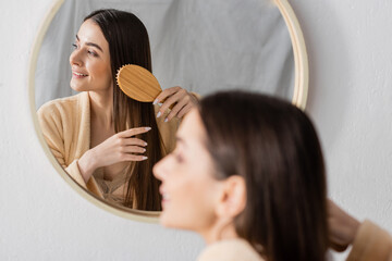 reflection of cheerful woman brushing shiny hair near mirror in bathroom.