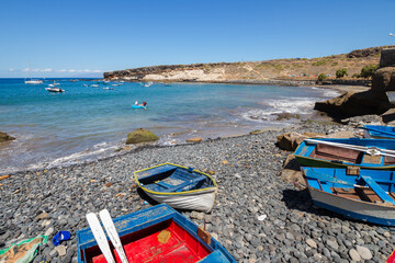 Several old fishing boats stranded on a pebble beach. Wooden boats of different colors, blue and white and red and white. Turquoise blue bay with yellow and brown hill in the background. La Caleta,