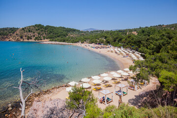 Peaceful summer day at the scenic Greek coastline, with turquoise bay waters and clear skies. Sunbeds, parasols and trees providing a tranquil holiday atmosphere.