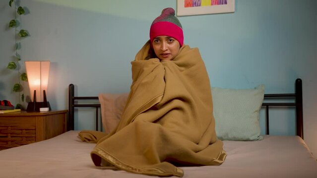 shivering Young girl full coverd with blanket and cap sitting on bed at during night - concept of chilling winter cold, climate change and influenza or fever