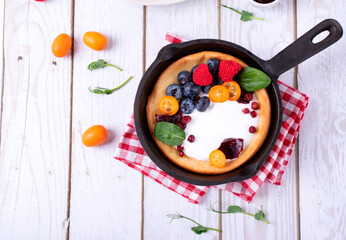 Dutch baby pancake served with custard, whipped cream and berries in cast iron pan on white wooden table. Top view