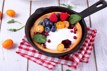 Dutch baby pancake served with custard, whipped cream and berries in cast iron pan on white wooden table