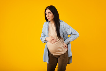 Young pregnant woman with contractions, feeling unwell while standing over yellow studio background