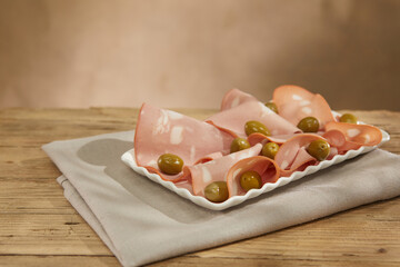 slices of mortadella on a white plate with olives. italian sausage on a wooden table with a napkin. serving meat delicacy. traditional snack under sunlight. rustic food.