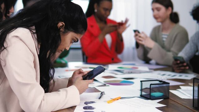 Pretty indian young female browsing internet using social media, sitting with diverse mixed race colleagues. Addicted students using smartphone. Communication problem. Youth Device addiction. School