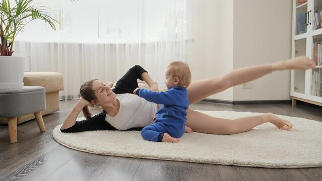 Cute baby boy watching young mother lying on floor and doing stretching and practising fitness. Family healthcare, active lifestyle, parenting and child development.