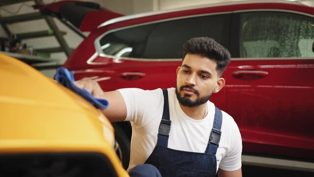 Bearded car wash worker man, wearing protective clothes uniform overalls, cleaning modern yellow luxury car with microfiber cloth. Car detailing or vehicle concept. Male worker holds the microfiber.