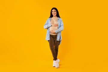 Fototapeta na wymiar Full length shot of happy pregnant woman touching belly and smiling, posing over yellow studio background