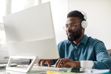 African american male employee in glasses and wireless headphones sitting at office desk and...