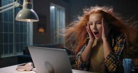 Positively surprised redhead caucasian girl looking at laptop, wing blowing her hair. Model reading...