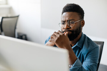 Thoughtful black man in eyeglasses stack with hard task, looking at computer screen, thinking of problem solution