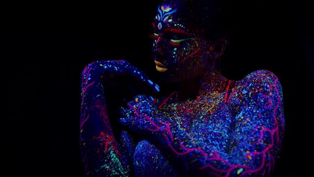 Young female performer with a painted face and fluorescent multicolor drips on his skin slowly touching herself on the shoulders in dance in UV light on a black background. Painted woman dancer