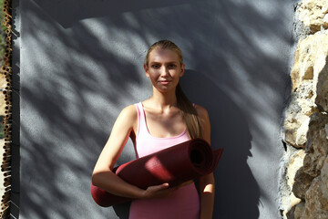 Young beautiful woman holding a rolled yoga mat. Blonde yogini leaning on the gray wall with patterns of sunlight and tree leaves shadows. Close up, copy space, background.