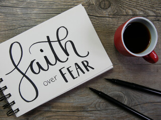 Top view of FAITH OVER FEAR lettering in notebook on wooden surface with coffee and pens
